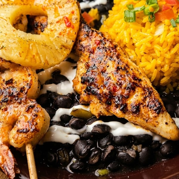 Grilled shrimp skewer, chicken, pineapple, yellow rice, and black beans with white sauce on a plate.