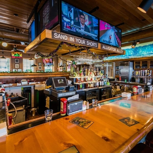 A rustic restaurant bar with a wooden counter, stools, liquor bottles, beer taps, and TVs showing sports.