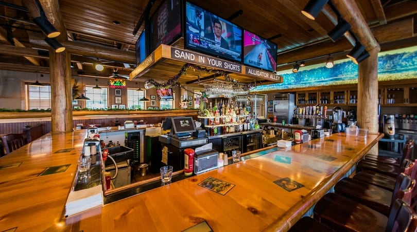 A rustic restaurant bar with a wooden counter, stools, liquor bottles, beer taps, and TVs showing sports.