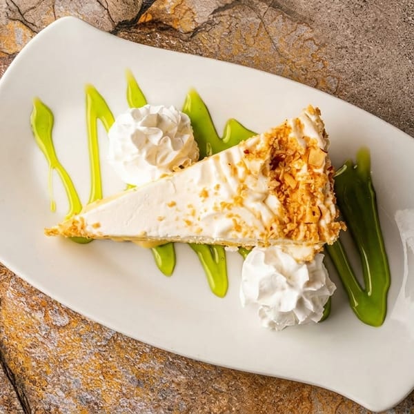 Slice of cheesecake with nuts, whipped cream, and green sauce on a white plate.