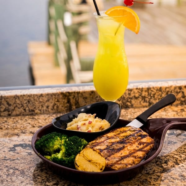 Grilled fish with broccoli, mashed potatoes, and a cocktail on a granite counter overlooking water.